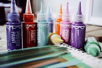 A collection of colorful poster paint bottles with pointed nozzle caps is arranged in a row. Vibrant hues include purple, red, teal, orange, pink, and yellow. In the foreground, a spattered notebook or paper displaying painted stripes in greens, blues, and browns is visible.