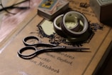 A pair of vintage-style scissors rests on a decorative journal with the words 'Memo, Daybook' and 'Notebook' on its cover. Beside the scissors are rolls of washi tape, and in the background, there are playing cards and other stationary items partially visible.