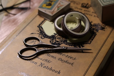 A pair of vintage-style scissors rests on a decorative journal with the words 'Memo, Daybook' and 'Notebook' on its cover. Beside the scissors are rolls of washi tape, and in the background, there are playing cards and other stationary items partially visible.