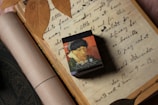 An open book or notebook displays handwritten script with cursive writing, accompanied by dried leaves placed on top of the pages. A small box featuring an artwork of a man wearing a hat and a coat is also present, partially covering the text.