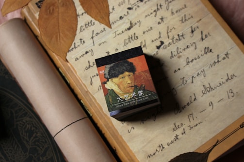 An open book or notebook displays handwritten script with cursive writing, accompanied by dried leaves placed on top of the pages. A small box featuring an artwork of a man wearing a hat and a coat is also present, partially covering the text.
