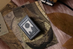 A collection of vintage-like astrology-themed items featuring an astrology book or card on top of celestial maps. The design includes circular and linear patterns reminiscent of old astronomical charts. A large, dried brown leaf is placed beside these items, contrasting with the soft textured surface beneath.