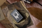 A collection of vintage-like astrology-themed items featuring an astrology book or card on top of celestial maps. The design includes circular and linear patterns reminiscent of old astronomical charts. A large, dried brown leaf is placed beside these items, contrasting with the soft textured surface beneath.