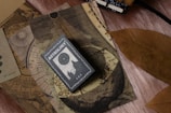 A collection of vintage-like astrology-themed items featuring an astrology book or card on top of celestial maps. The design includes circular and linear patterns reminiscent of old astronomical charts. A large, dried brown leaf is placed beside these items, contrasting with the soft textured surface beneath.