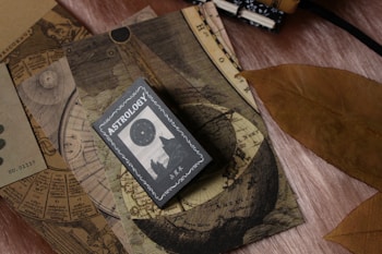 A collection of vintage-like astrology-themed items featuring an astrology book or card on top of celestial maps. The design includes circular and linear patterns reminiscent of old astronomical charts. A large, dried brown leaf is placed beside these items, contrasting with the soft textured surface beneath.