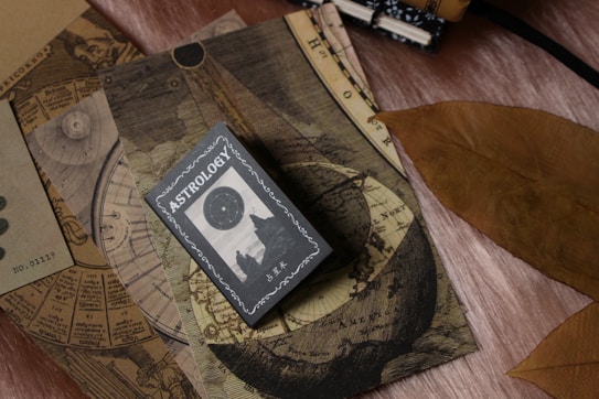 A collection of vintage-like astrology-themed items featuring an astrology book or card on top of celestial maps. The design includes circular and linear patterns reminiscent of old astronomical charts. A large, dried brown leaf is placed beside these items, contrasting with the soft textured surface beneath.
