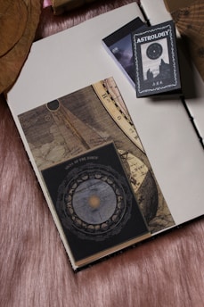 An open book with a vintage celestial and zodiac theme rests on a soft, textured fabric background. Next to the book is a deck with the label 'Astrology' featuring an image of the night sky and silhouettes of two figures. The book pages show intricate designs with celestial maps and astrology symbols.