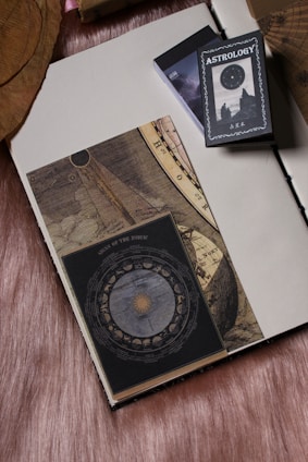 An open book with a vintage celestial and zodiac theme rests on a soft, textured fabric background. Next to the book is a deck with the label 'Astrology' featuring an image of the night sky and silhouettes of two figures. The book pages show intricate designs with celestial maps and astrology symbols.