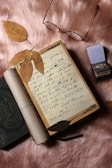 An open journal with handwritten text, surrounded by dried leaves, an old rolled parchment, a pair of reading glasses, a deck of playing cards, and a closed book with a decorative cover, all laid on a soft pink textured surface.