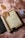 An open journal with handwritten text, surrounded by dried leaves, an old rolled parchment, a pair of reading glasses, a deck of playing cards, and a closed book with a decorative cover, all laid on a soft pink textured surface.
