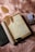An open journal with handwritten text, surrounded by dried leaves, an old rolled parchment, a pair of reading glasses, a deck of playing cards, and a closed book with a decorative cover, all laid on a soft pink textured surface.