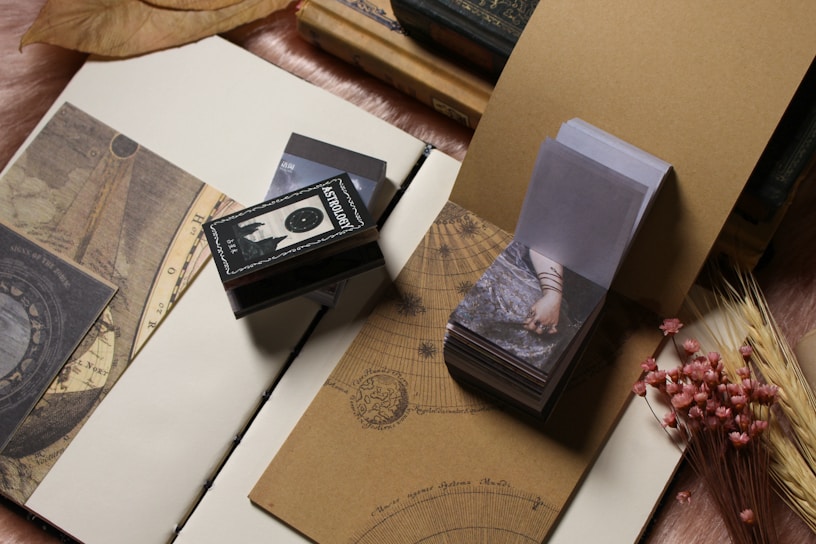 An array of vintage and artistic items is displayed, including books with old covers and zodiac sketches, an astrology-themed matchbox, photographic images featuring an arm adorned with bracelets, and pink dried flowers arranged near wheat stalks. The setup conveys a sense of nostalgia and mystique, with items carefully laid on an open notebook.