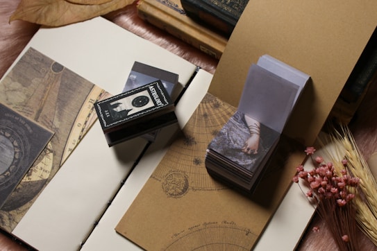 An array of vintage and artistic items is displayed, including books with old covers and zodiac sketches, an astrology-themed matchbox, photographic images featuring an arm adorned with bracelets, and pink dried flowers arranged near wheat stalks. The setup conveys a sense of nostalgia and mystique, with items carefully laid on an open notebook.