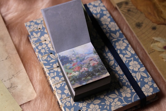 A small notebook with a blue floral cover is lying on a soft, textured surface. An open flipbook or notepad with a colorful illustration of a garden is placed on top, partially covering the notebook.