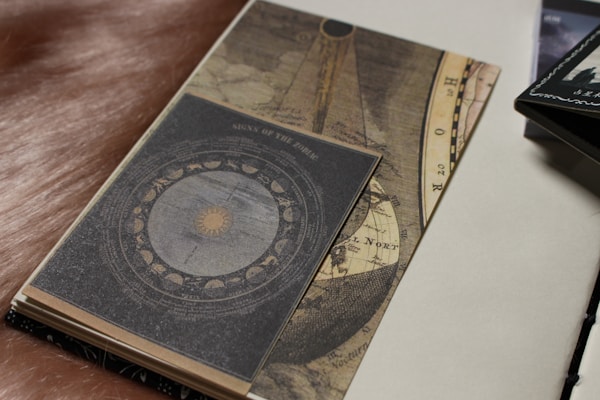 A vintage-style notebook or journal lies open, displaying an intricate design featuring astrological symbols and a zodiac wheel. The cover has a worn, textured look with earthy tones and celestial motifs. Next to it, part of another book or paper is visible.
