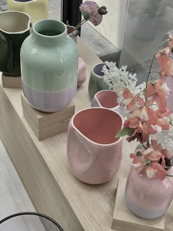 Various ceramic vases in pastel colors, including shades of mint green, light pink, yellow, and lilac, are arranged on wooden platforms. Some vases hold pink and white flowers, adding a soft, decorative touch to the display. The scene is set near a large window, allowing natural light to enhance the colors and textures of the ceramics.