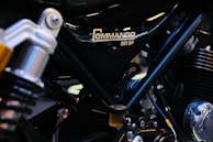 A close-up view of a motorcycle's engine area, showcasing the brand name 'Commando 961 SP' on the black metal of the bike. The image highlights the intricate details and components of the motorcycle, including the suspension and other metallic parts, reflecting a polished and well-maintained machine.