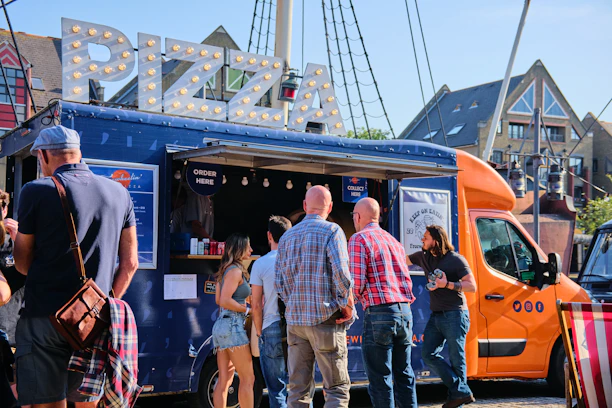 Happy customers enjoying their pizzas around the cozy trailer at a lively outdoor event.