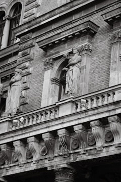 A detailed façade of a historic building featuring classical architecture. Two stone statues of draped figures stand in alcoves, one on each side, amidst ornate decorative elements such as columns and arches. The building is constructed with brick and stone, and there are intricate carvings and moldings around the windows and along the balustrade.