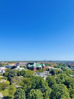A clear, sunny day showcases a landscape with lush green trees and a large, impressive building with a green roof and red brick facade. Surrounding the building are parking lots and urban infrastructure. The horizon reveals more buildings and a flat terrain extending into the distance beneath a vibrant blue sky.