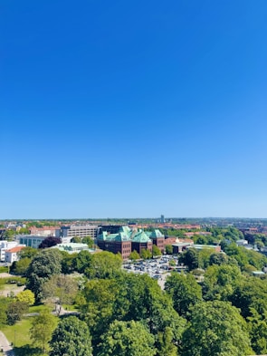 A clear, sunny day showcases a landscape with lush green trees and a large, impressive building with a green roof and red brick facade. Surrounding the building are parking lots and urban infrastructure. The horizon reveals more buildings and a flat terrain extending into the distance beneath a vibrant blue sky.