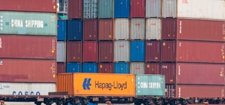 Multiple stacked shipping containers in various colors, including red, green, blue, and orange. The containers are branded with names such as Hapag-Lloyd and China Shipping, and they are stacked on a freight train.