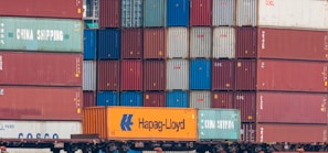 Multiple stacked shipping containers in various colors, including red, green, blue, and orange. The containers are branded with names such as Hapag-Lloyd and China Shipping, and they are stacked on a freight train.