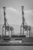 A monochrome image features two large port cranes towering above a row of shipping containers at a dockside. The cranes appear metallic and industrial, with a backdrop of distant hills and cloudy skies. Below the cranes and containers, there is a structure with a sloped roof and rows of windows, possibly a warehouse.