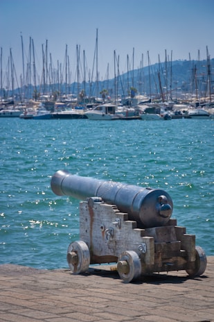 An old cannon rests on a brick-paved platform by a body of water, with numerous sailboats moored in the marina in the background. The scene includes a distant view of hills under a clear, blue sky.