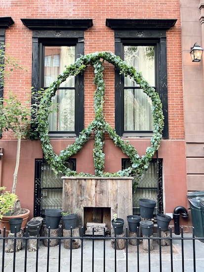 A large peace sign made of green foliage is mounted on the exterior of a red brick building with two black-framed windows. Below the peace sign is a rustic wooden structure surrounded by a black fence and multiple black plant pots. A tree with a terracotta pot and a lit wall-mounted lantern add to the urban setting.