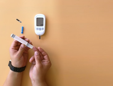 A person is preparing to check their blood sugar with a glucose meter. The hands are holding a lancet device near a finger, and a glucose meter with test strips is on the table.