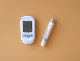 A blood glucose meter displaying a reading of 103 mg/dL is next to a lancing device on a solid light brown background. The meter and device are white with grey accents.