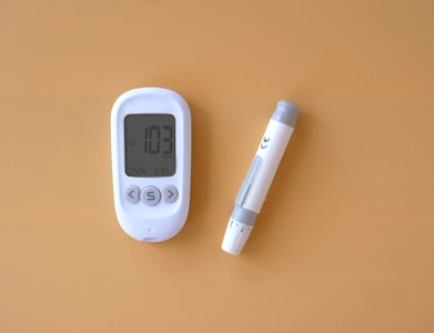 A blood glucose meter displaying a reading of 103 mg/dL is next to a lancing device on a solid light brown background. The meter and device are white with grey accents.