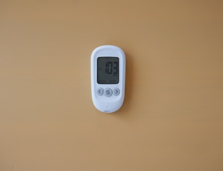 A white digital thermometer or thermostat is mounted on a plain, light brown or tan wall. The display shows '103' with additional smaller text below.