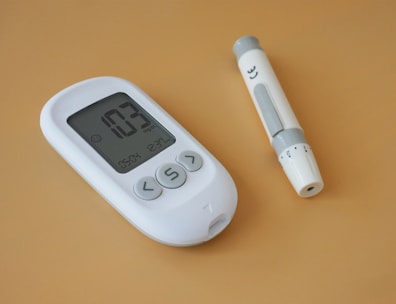 A white digital blood glucose meter displaying a reading of 103 mg/dL is placed next to a white lancet device on a light brown surface.