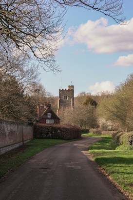 A winding country road bordered by green grass and hedges leads to a rustic brick house with a steep roof. Behind the house, a stone church tower rises against a backdrop of bare tree branches and a blue sky scattered with white clouds.