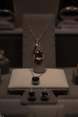 A display of elegant jewelry including a central necklace with a large gemstone, earrings, and other jewels arranged on a soft, neutral-colored surface under soft lighting.
