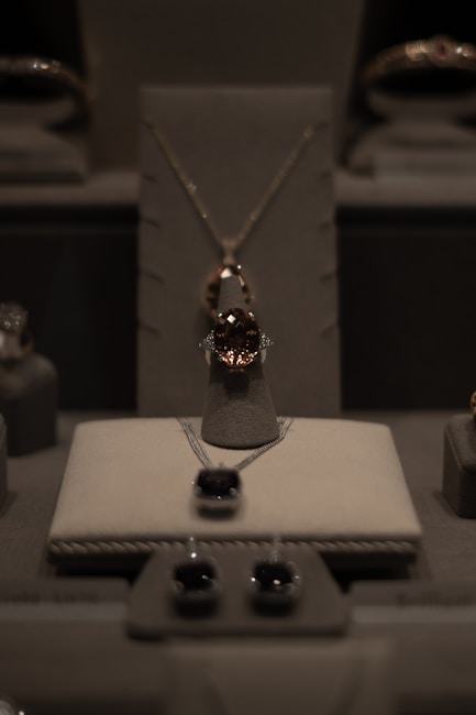 A display of elegant jewelry including a central necklace with a large gemstone, earrings, and other jewels arranged on a soft, neutral-colored surface under soft lighting.