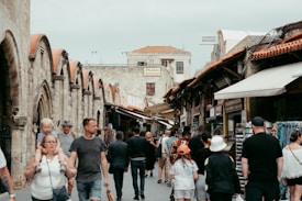A bustling street market filled with people walking along a narrow path lined by historic stone buildings with red-tiled roofs. Stalls and shops display various goods, and the atmosphere suggests a casual, lively environment. Some shoppers are casually dressed, and a child is being carried by an adult.