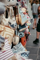 A variety of colorful bags are displayed, hanging neatly on racks in a street market setting. The bags feature different designs, such as stripes, patterns, and woven textures.