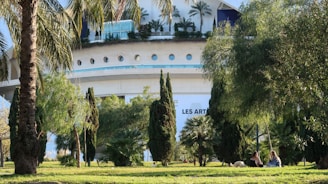 A modern architectural building with circular windows and lush greenery in the foreground. Palm trees and various bushes are scattered around the scene, with two people sitting on the grass in the shade, accompanied by a dog. The building has the words 'LES ARTS' visible on it.