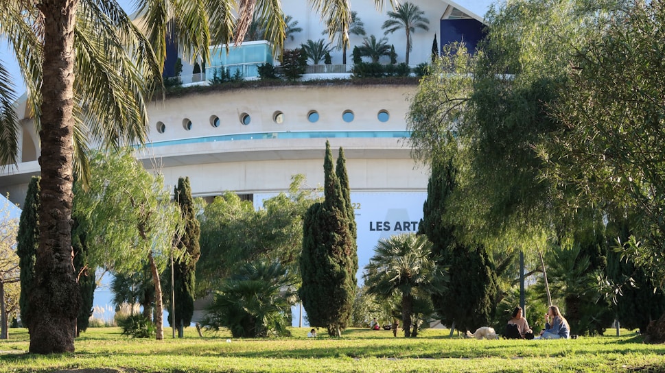 A modern architectural building with circular windows and lush greenery in the foreground. Palm trees and various bushes are scattered around the scene, with two people sitting on the grass in the shade, accompanied by a dog. The building has the words 'LES ARTS' visible on it.