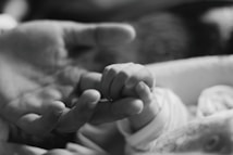 A small baby's hand gently gripping an adult's finger, symbolizing a tender moment of connection and care. The image is in black and white, highlighting the textures and wrinkles of the skin.