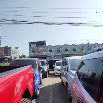 A crowded parking area features several cars and trucks parked closely together, with a row of low-rise commercial buildings visible in the background. The buildings have a modern design with green and gray accents. Overhead, numerous electrical wires are stretched across the scene.