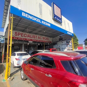 A car service workshop with prominent signage advertising its specialization in automotive air conditioning. Several cars are parked in front, including a red SUV and a white sedan. The building has large signs in Indonesian and a blue banner with the text 'BENGKEL RESMI' and 'AGAM AC'.