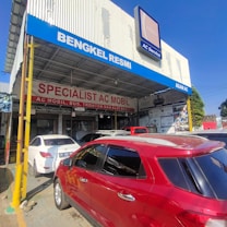 A car service workshop with prominent signage advertising its specialization in automotive air conditioning. Several cars are parked in front, including a red SUV and a white sedan. The building has large signs in Indonesian and a blue banner with the text 'BENGKEL RESMI' and 'AGAM AC'.