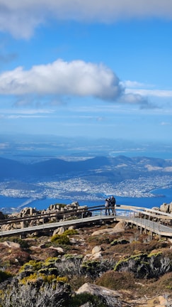 A scenic landscape featuring a rocky hillside with sparse vegetation in the foreground. A wooden boardwalk winds its way through the rocks, where two individuals are standing and taking a photograph. In the background, a sprawling cityscape and a large body of water are visible, with a bridge crossing the water. The sky above is blue and dotted with fluffy white clouds.