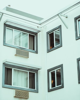 A section of a modern building exterior featuring several square windows framed in dark gray. The walls are painted a light cream color, and air conditioning units are visible below the windows. The image appears to be taken at an upward angle, focusing on the corner of the building.