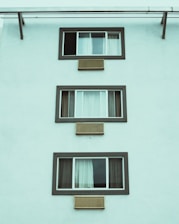 A minimalist facade of a building featuring three rectangular windows, aligned vertically. Each window is framed with dark trim and accompanied by horizontal air conditioning vents below. The curtain arrangements vary across the windows, with the first window partially open.