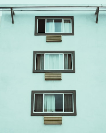 A minimalist facade of a building featuring three rectangular windows, aligned vertically. Each window is framed with dark trim and accompanied by horizontal air conditioning vents below. The curtain arrangements vary across the windows, with the first window partially open.